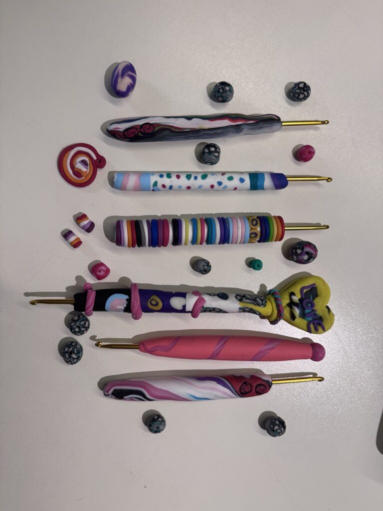 Verschiedene bunte Häkelnadeln mit Griffen aus Fimo, in den Farben verschiedener Pride-Flaggen, umgeben von Fimo-Bällen